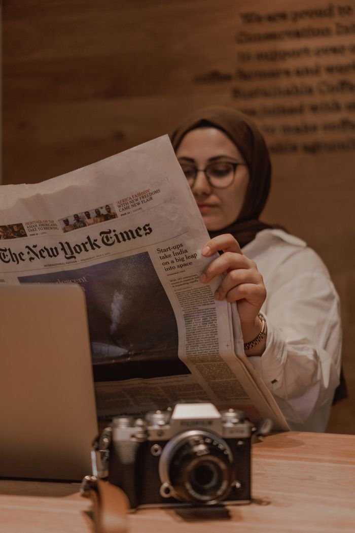 Young woman in a hijab reads The New York Times in a warm café atmosphere, showcasing concentration and relaxation.